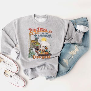 May include: A gray sweatshirt with a vintage-style graphic of Mickey Mouse and other Disney characters on a pirate ship. The text "Pirates of the Caribbean Disneyland" is printed on the sweatshirt.