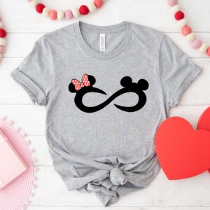 May include: Grey t-shirt featuring a black infinity symbol design. One side of the infinity symbol has a red bow, and the other side has a black Mickey Mouse ear.