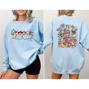 Op de afbeelding: Lichtblauw sweatshirt met een feestelijk Disney-ontwerp. De voorkant toont Disney-personages in feestelijke kleding met lichtslingers. De achterkant toont een kleurrijke Walt Disney World-afbeelding met personages en cadeaus. Een comfortabele top met lange mouwen.