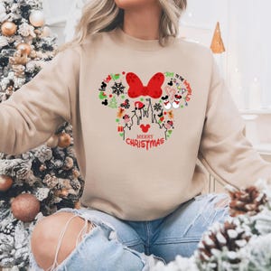 Pu&ograve; includere: Felpa beige con un design natalizio a forma di Minnie Mouse. Il design include un fiocco rosso, fiocchi di neve, teste di Topolino e il testo "Merry Christmas" e "Joy to the World."