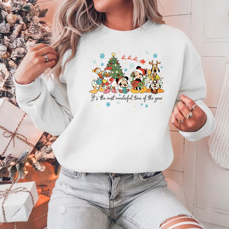 May include: White sweatshirt featuring a colorful graphic of Disney characters in Christmas attire, including Mickey Mouse, Donald Duck, and Pluto, with a Christmas tree and snowflakes. The text "It's the most wonderful time of the year" is printed below the graphic.