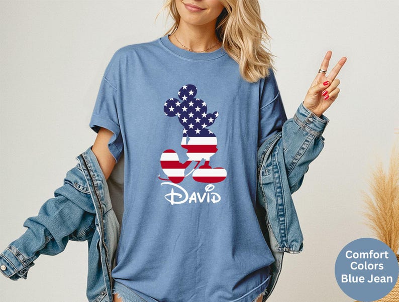 May include: Blue jean-colored t-shirt featuring a Mickey Mouse silhouette filled with a stars and stripes design. The name "David" is written in white script below the character. The shirt is part of the Comfort Colors collection.
