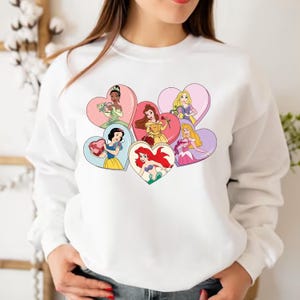 Pode incluir: Camisola branca com um gr&aacute;fico colorido de seis princesas Disney segurando cora&ccedil;&otilde;es. Cada princesa est&aacute; em uma moldura diferente em forma de cora&ccedil;&atilde;o.