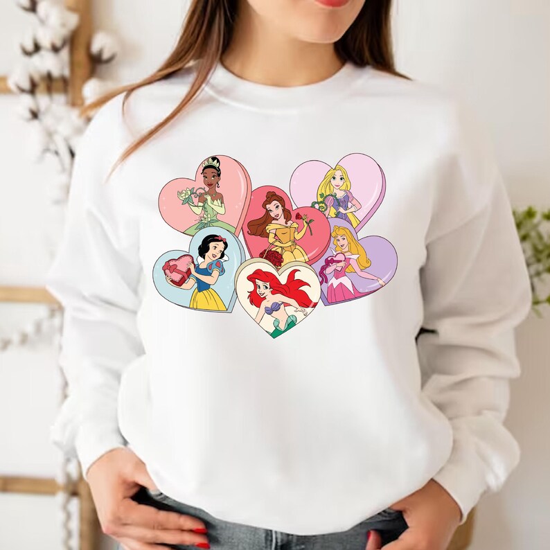 Pode incluir: Camisola branca com um gr&aacute;fico colorido de seis princesas Disney segurando cora&ccedil;&otilde;es. Cada princesa est&aacute; em uma moldura diferente em forma de cora&ccedil;&atilde;o.
