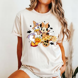 May include: A cream-colored t-shirt with a graphic of Minnie Mouse and Daisy Duck dressed in Halloween costumes. Minnie Mouse is wearing a witch's hat and holding a pumpkin. Daisy Duck is wearing a witch's hat and holding a pumpkin. The graphic is in a cartoon style.