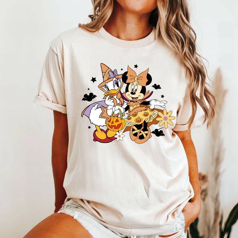 May include: A cream-colored t-shirt with a graphic of Minnie Mouse and Daisy Duck dressed in Halloween costumes. Minnie Mouse is wearing a witch's hat and holding a pumpkin. Daisy Duck is wearing a witch's hat and holding a pumpkin. The graphic is in a cartoon style.