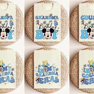 Op de afbeelding: Cr&egrave;mekleurige T-shirts met blauwe tekst en Mickey Mouse-afbeeldingen. De shirts hebben zinnen als "Opa van de jarige", "Oma van de jarige" en "Neef van de jarige". Andere shirts zeggen "In mijn opa-tijdperk", "In mijn oma-tijdperk" en "In mijn neef-tijdperk".