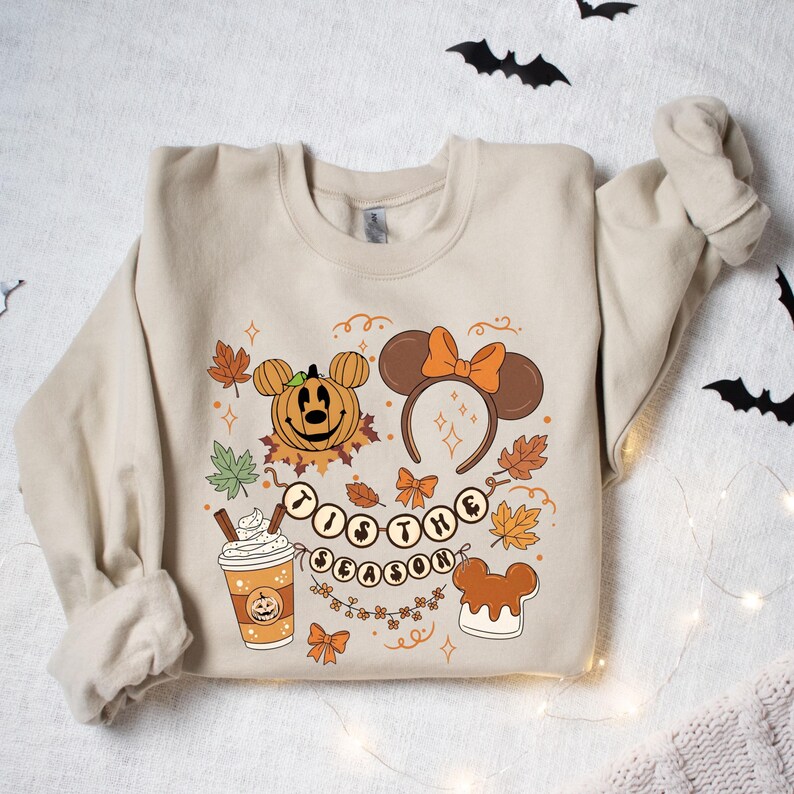 May include: Beige sweatshirt with a Halloween-themed graphic. The design includes a pumpkin, Mickey Mouse ears headband, fall leaves, and the phrase "'Tis the Season." A coffee cup and a Mickey Mouse-shaped dessert are also featured.