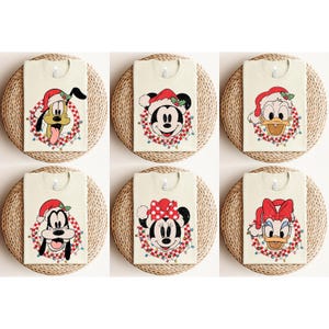 May include: Six white t-shirts with different Christmas-themed Disney character designs. The characters include Mickey Mouse, Minnie Mouse, Donald Duck, Daisy Duck, Pluto, and Goofy. Each character is wearing a Santa hat and is surrounded by a red and white wreath with Christmas lights.