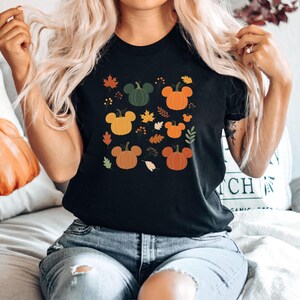 May include: Black t-shirt featuring a fall-themed design. The design includes various pumpkins in orange, yellow, and green, along with autumn leaves. The pumpkins have Mickey Mouse ear shapes. The shirt is a crew neck.