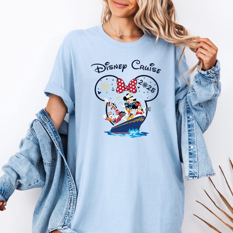 May include: Light blue t-shirt with a Disney Cruise graphic. The graphic features Mickey and Minnie Mouse, along with other Disney characters, on a cruise ship. The words "Disney Cruise 2025" are printed above the ship in a Mickey Mouse head shape.