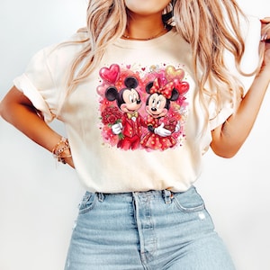 May include: Cream-colored t-shirt featuring a Valentine's Day graphic of Mickey and Minnie Mouse. Mickey is in a red suit, holding roses, and Minnie is in a red polka-dot dress, holding a heart-shaped gift. Balloons and hearts surround the couple.