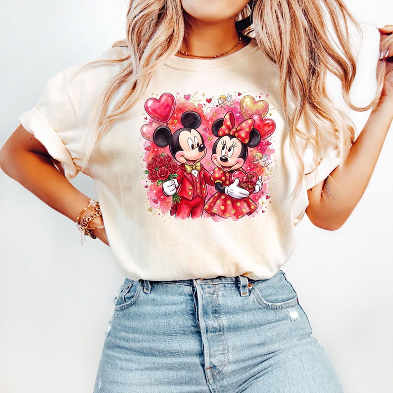 May include: Cream-colored t-shirt featuring a Valentine's Day graphic of Mickey and Minnie Mouse. Mickey is in a red suit, holding roses, and Minnie is in a red polka-dot dress, holding a heart-shaped gift. Balloons and hearts surround the couple.