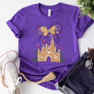 May include: Purple t-shirt with a graphic of a cartoon castle with a Mickey Mouse head on the front. The castle is surrounded by fireworks and a bow with polka dots.