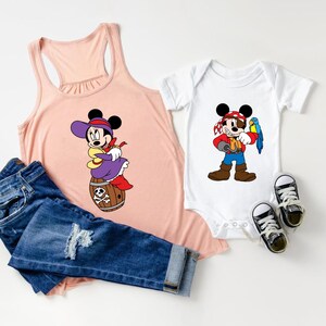 May include: A peach-colored tank top and a white baby onesie, both featuring cartoon pirate-themed characters. The tank top is paired with blue jeans. Two pairs of black and white sneakers are also in the image.