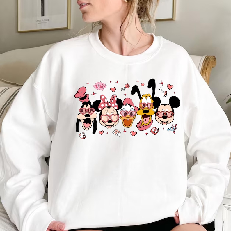 Op de afbeelding: Witte trui met een grafische print van Mickey Mouse, Minnie Mouse, Donald Duck en Goofy die hartvormige zonnebrillen dragen. De grafiek bevat ook harten, sterren en het woord "LOVE!"