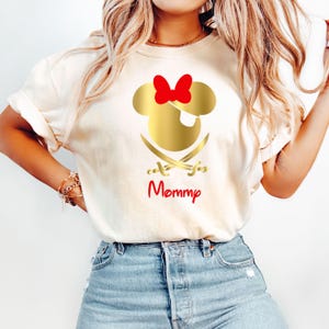 May include: White t-shirt with a gold Mickey Mouse head with a red bow and an eye patch. Two gold crossed swords are below the head. The text "Mommy" is written in red below the swords.