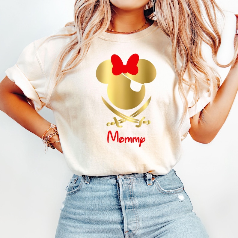 May include: White t-shirt with a gold Mickey Mouse head with a red bow and an eye patch. Two gold crossed swords are below the head. The text "Mommy" is written in red below the swords.