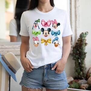 May include: White t-shirt with a colorful design featuring Mickey Mouse, Minnie Mouse, Donald Duck, Daisy Duck, and Goofy. Each character is wearing a different colored bow.