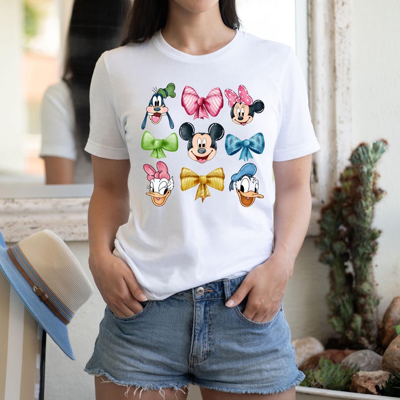May include: White t-shirt with a colorful design featuring Mickey Mouse, Minnie Mouse, Donald Duck, Daisy Duck, and Goofy. Each character is wearing a different colored bow.