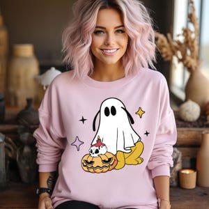 May include: A light pink sweatshirt featuring a cartoon ghost design with a jack-o'-lantern and stars. The ghost is white with black eyes and ears. The jack-o'-lantern is orange with a skull and candle. The sweatshirt is a crew neck style.