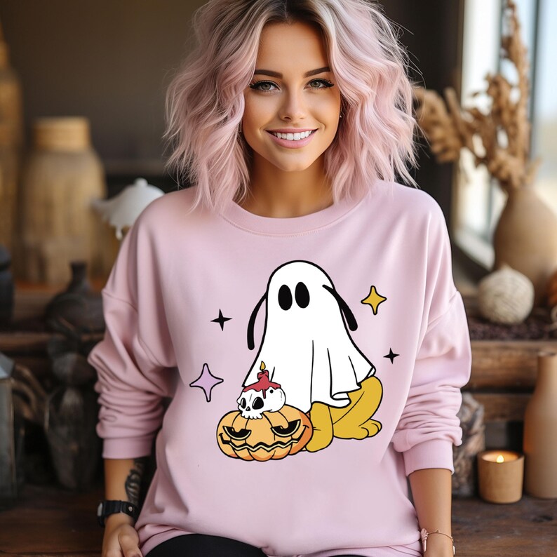 May include: A light pink sweatshirt featuring a cartoon ghost design with a jack-o'-lantern and stars. The ghost is white with black eyes and ears. The jack-o'-lantern is orange with a skull and candle. The sweatshirt is a crew neck style.