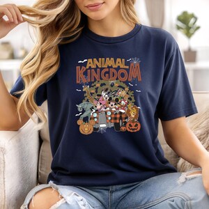 Op de afbeelding: Marineblauw T-shirt met een grafische afbeelding met de tekst "ANIMAL KINGDOM" en verschillende stripfiguren in een jeep. Het ontwerp bevat pompoenen en spinnenwebben, wat wijst op een Halloween-thema.