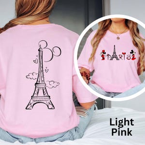 K&ouml;nnte beinhalten: Hellrosa T-Shirt mit einem schwarzen Eiffelturm-Design, Mickey Mouse Ohren und Herzen auf dem R&uuml;cken. Auf der Vorderseite steht das Wort "PARIS" mit Minnie- und Mickey-Mouse-Figuren. Das T-Shirt hat eine hellrosa Farbe.