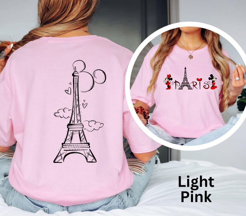 K&ouml;nnte beinhalten: Hellrosa T-Shirt mit einem schwarzen Eiffelturm-Design, Mickey Mouse Ohren und Herzen auf dem R&uuml;cken. Auf der Vorderseite steht das Wort "PARIS" mit Minnie- und Mickey-Mouse-Figuren. Das T-Shirt hat eine hellrosa Farbe.