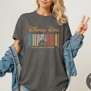 May include: Dark grey t-shirt with the text "Disney Dad" in gold. Below the text is a barcode design with a castle and Mickey Mouse silhouette. The text "SCAN FOR PAYMENT" is below the barcode.