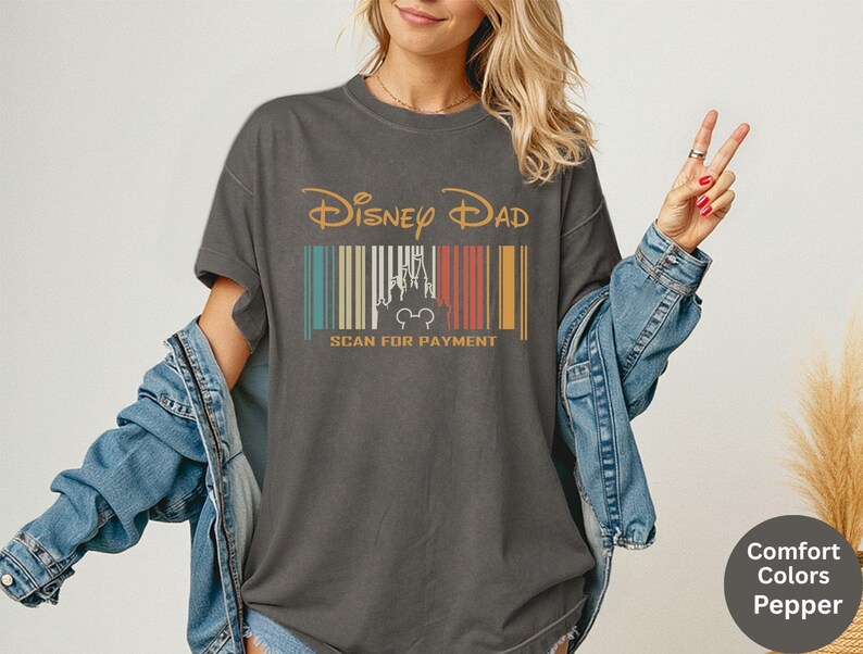 May include: Dark grey t-shirt with the text "Disney Dad" in gold. Below the text is a barcode design with a castle and Mickey Mouse silhouette. The text "SCAN FOR PAYMENT" is below the barcode.