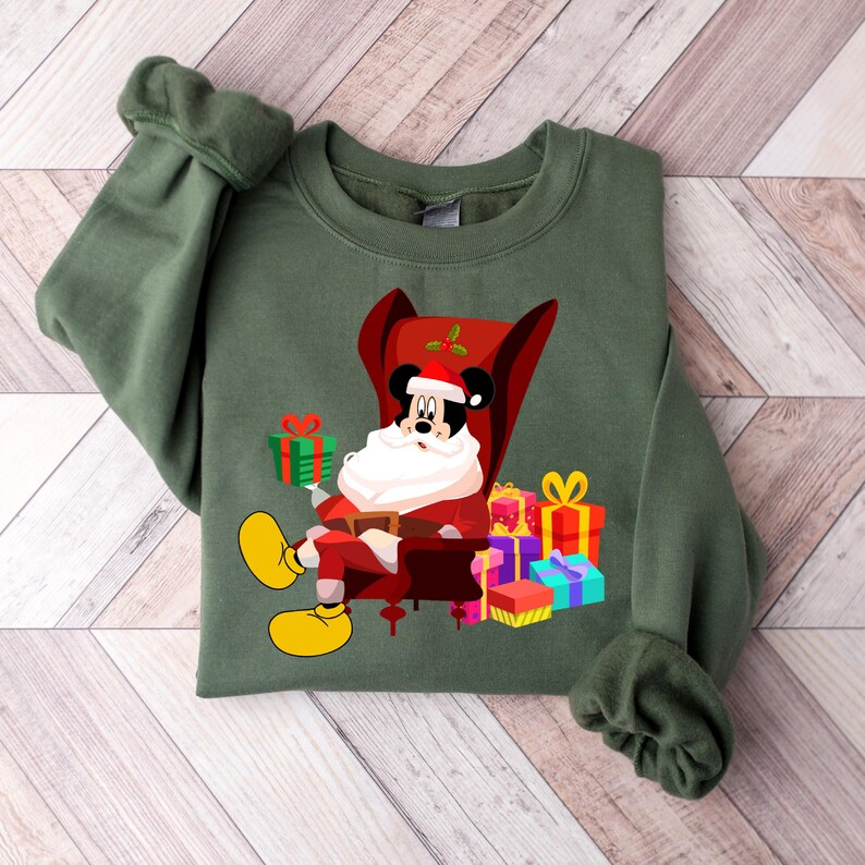 Op de afbeelding: Groen sweatshirt met een feestelijk Mickey Mouse-ontwerp. Mickey, verkleed als kerstman, zit in een rode stoel, omringd door ingepakte cadeaus. Het ontwerp bevat een kerstmuts, baard en gele schoenen, ideaal voor de feestdagen.