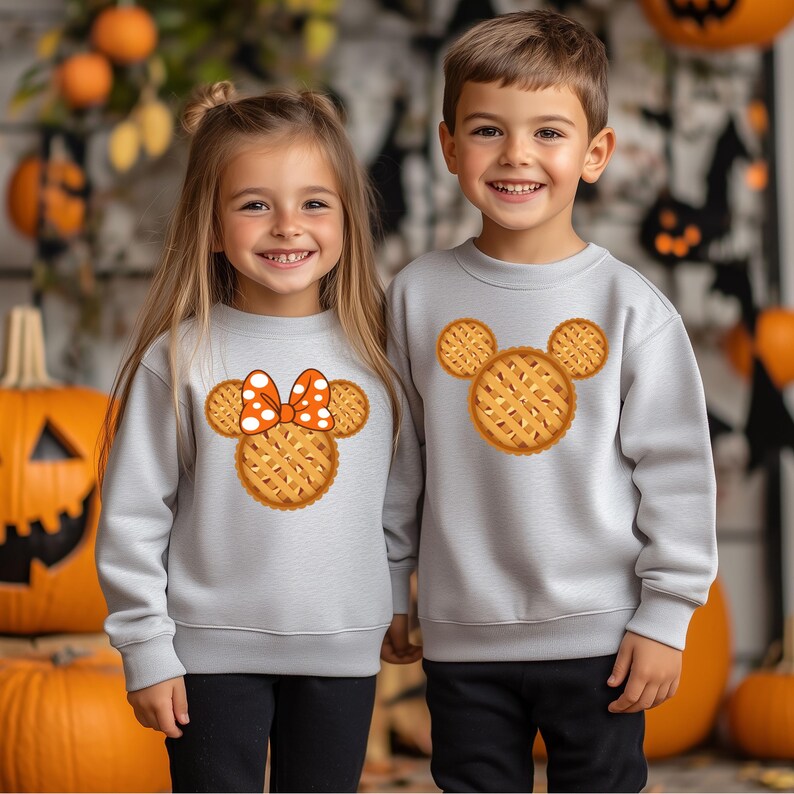 K&ouml;nnte beinhalten: Zwei Kinder tragen hellgraue Sweatshirts mit einem Kuchen-f&ouml;rmigen Design. Ein Sweatshirt hat eine Schleife mit orangefarbenen Punkten. Das andere hat ein Kuchen-f&ouml;rmiges Design. K&uuml;rbisse und Herbstdekorationen sind im Hintergrund.