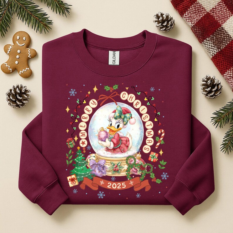 Pode incluir: Camisola bord&ocirc; com um design de globo de neve com a Margarida segurando um enfeite rosa. As palavras "Merry Christmas" e o ano "2025" s&atilde;o exibidos. Roupas festivas de Natal.