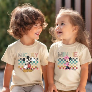 May include: Two children wearing matching t-shirts with a retro design. The boy's shirt features Mickey Mouse and the text "MICKEY" in a colorful, layered font. The girl's shirt features Minnie Mouse and the text "MINNIE" in the same font.