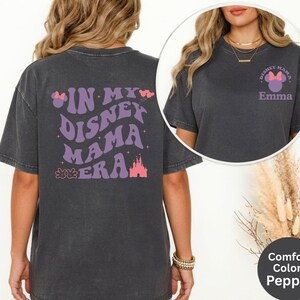 Puede incluir: Una camiseta gris oscuro con el texto "IN MY DISNEY MAMA ERA" en morado, junto con un castillo y un lazo gr&aacute;fico. La parte delantera de la camiseta tiene un peque&ntilde;o gr&aacute;fico que dice "Disney Mama" con un lazo y el nombre "Emma".