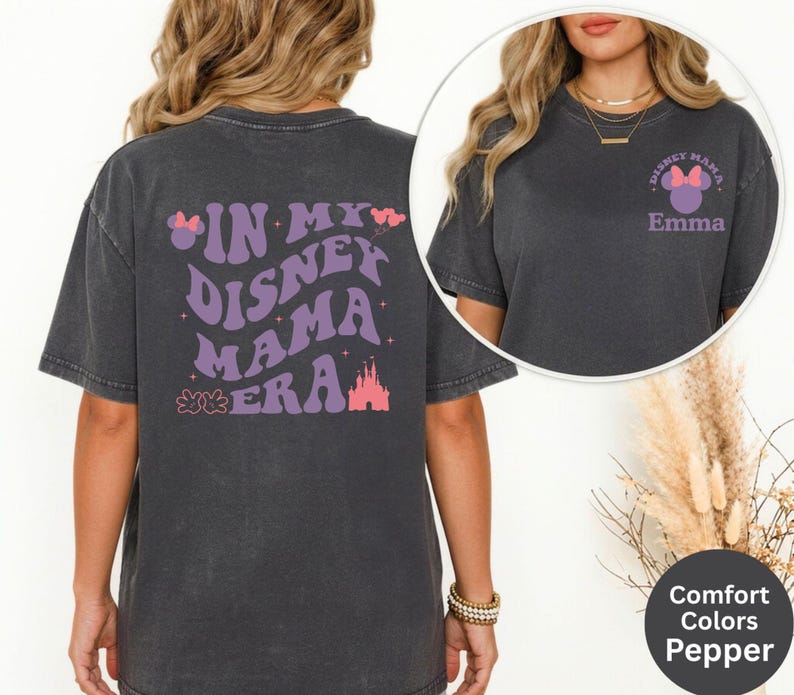 Puede incluir: Una camiseta gris oscuro con el texto "IN MY DISNEY MAMA ERA" en morado, junto con un castillo y un lazo gr&aacute;fico. La parte delantera de la camiseta tiene un peque&ntilde;o gr&aacute;fico que dice "Disney Mama" con un lazo y el nombre "Emma".