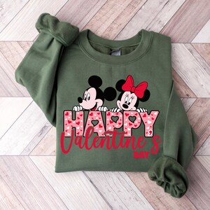 May include: Green sweatshirt featuring a graphic of Mickey Mouse and Minnie Mouse peeking over the words "Happy Valentine's Day" with pink hearts in the letters.