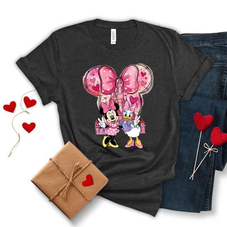 May include: Dark gray t-shirt featuring Minnie Mouse and Daisy Duck with a pink bow and castle design. The graphic includes pink hearts and the characters are giving peace signs. The shirt is paired with blue jeans.