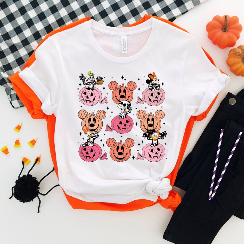 May include: White t-shirt with a graphic print of Mickey Mouse and friends dressed as pumpkins. The design features a variety of characters, including Mickey Mouse, Minnie Mouse, Donald Duck, and Goofy.