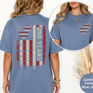May include: Blue jean-colored t-shirt with a distressed American flag design. The back features a large flag graphic with the text "250 YEARS". The front has a smaller flag design. The shirt is a comfort colors style.