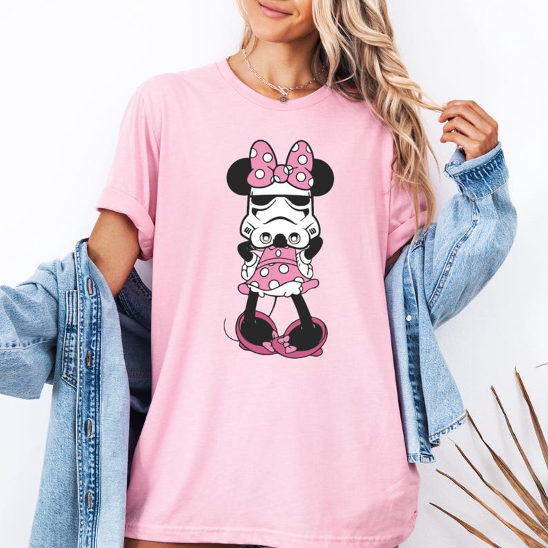 May include: Pink t-shirt featuring a cartoon mashup of Minnie Mouse and a Stormtrooper. The design includes a polka-dot bow, dress, and shoes. The graphic is in black, white, and pink. The shirt is a short-sleeved crew neck.