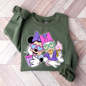 Pode incluir: Uma camisa de frio verde com uma estampa gr&aacute;fica de dois personagens de desenho animado usando &oacute;culos de sol em forma de cora&ccedil;&atilde;o. Os personagens est&atilde;o segurando uma bebida e um cone de sorvete.