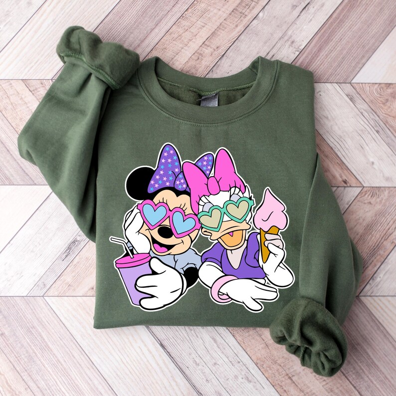 Pode incluir: Uma camisa de frio verde com uma estampa gr&aacute;fica de dois personagens de desenho animado usando &oacute;culos de sol em forma de cora&ccedil;&atilde;o. Os personagens est&atilde;o segurando uma bebida e um cone de sorvete.