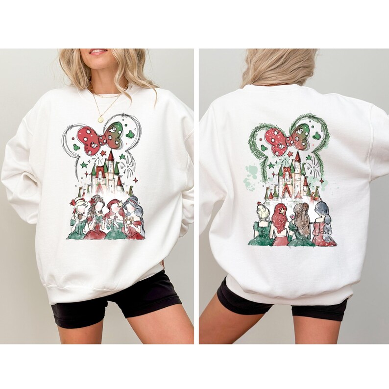 May include: White sweatshirt featuring a watercolor design of a castle, fireworks, and several princesses. The design is framed by a Mickey Mouse ear shape with a red and green bow. The sweatshirt is worn by a person.