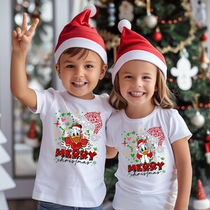 May include: Two children wearing white t-shirts with a red and green Christmas design featuring a cartoon duck. The shirts say "Merry Christmas". The children are wearing red Santa hats.
