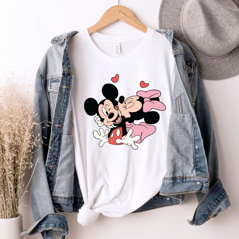 May include: White t-shirt featuring a graphic of Mickey Mouse kissing Minnie Mouse, with red heart accents. The cartoon characters are in black, red, and pink. The shirt is displayed on a wooden hanger, with a denim jacket and a hat in the background.