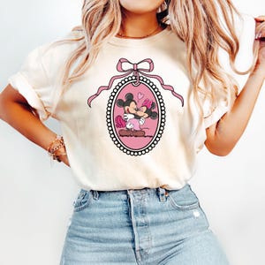 K&ouml;nnte beinhalten: Ein wei&szlig;es T-Shirt mit einer rosa Schleife und einem rosa ovalen Rahmen, der Mickey Mouse und Minnie Mouse beim K&uuml;ssen zeigt. Der Rahmen ist mit wei&szlig;en Perlen verziert.
