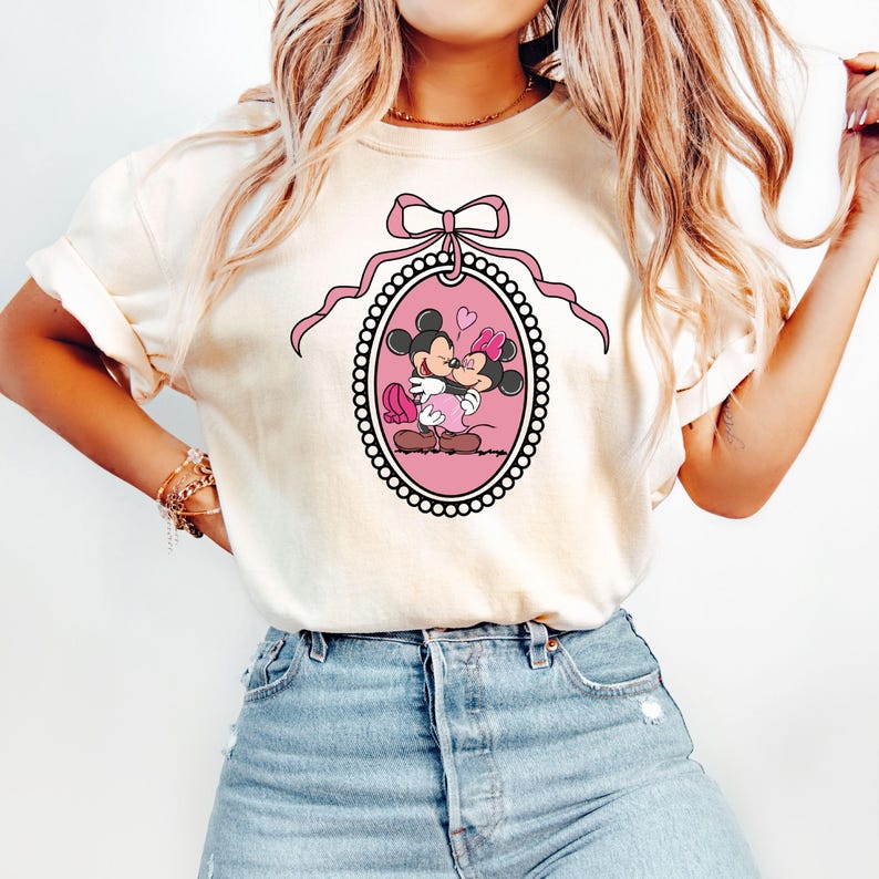 K&ouml;nnte beinhalten: Ein wei&szlig;es T-Shirt mit einer rosa Schleife und einem rosa ovalen Rahmen, der Mickey Mouse und Minnie Mouse beim K&uuml;ssen zeigt. Der Rahmen ist mit wei&szlig;en Perlen verziert.
