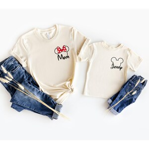 May include: Two cream-colored t-shirts with black text and graphics. One shirt has a Minnie Mouse bow and the word "Mom". The other shirt has a Mickey Mouse head outline and the name "Jerry". Blue jeans are next to the shirts.